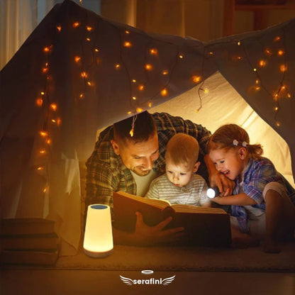 Um pai brincando de "barraca" de lençol no quarto com seus dois filhos e lendo um livro junto com eles, com o auxílio da luminária acesa.