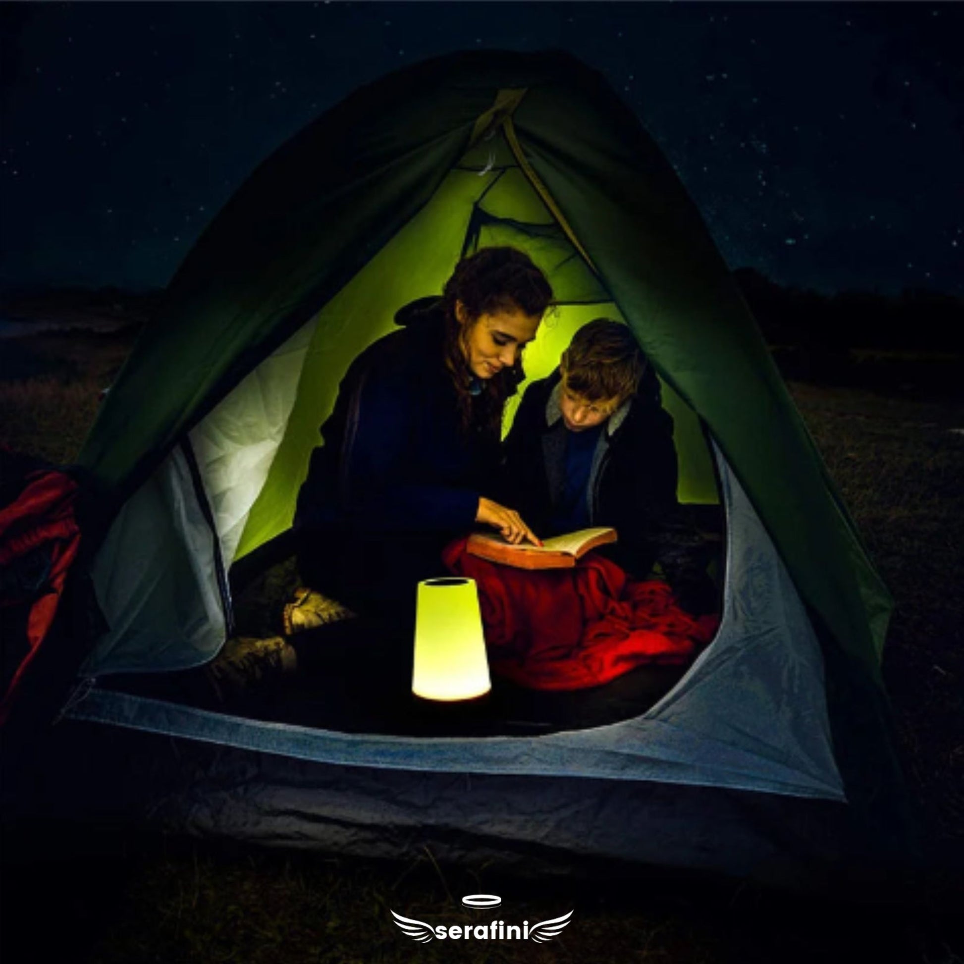 Uma mãe e seu filho, dentro de uma barraca em ambiente externo, lendo um livro juntos com a luminária portátil dentro da barraca.