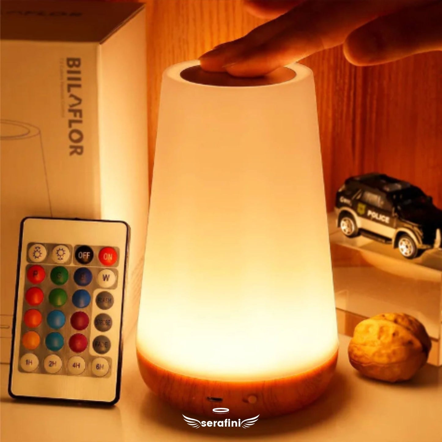 Luminária acesa com luz branca, uma mão sobre o sensor de toque. Ao lafo o controle remoto e itens de decoração em uma prateleira.