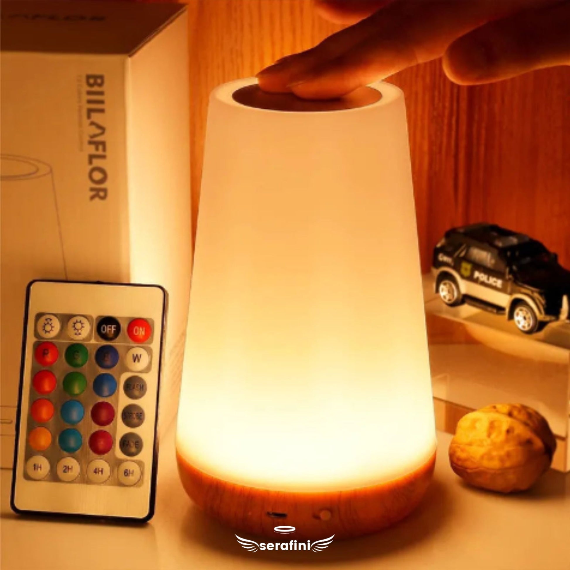 Luminária acesa com luz branca, uma mão sobre o sensor de toque. Ao lafo o controle remoto e itens de decoração em uma prateleira.