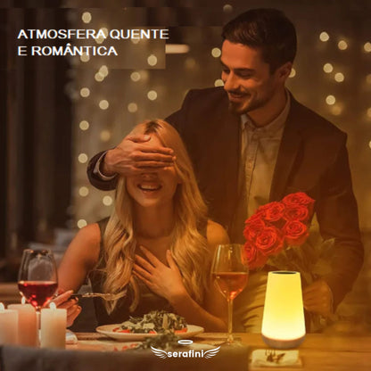 Uma cena romântica de um jantar, com uma linda mesa posta, uma taça de vinho tinto e a luminária delicada ao lado. Uma mulher está sentada à mesa com o garfo em mãos e um homem chega de surpresa e cobre seus olhos, para surpreendê-la com um buquê de rosas vermelhas.